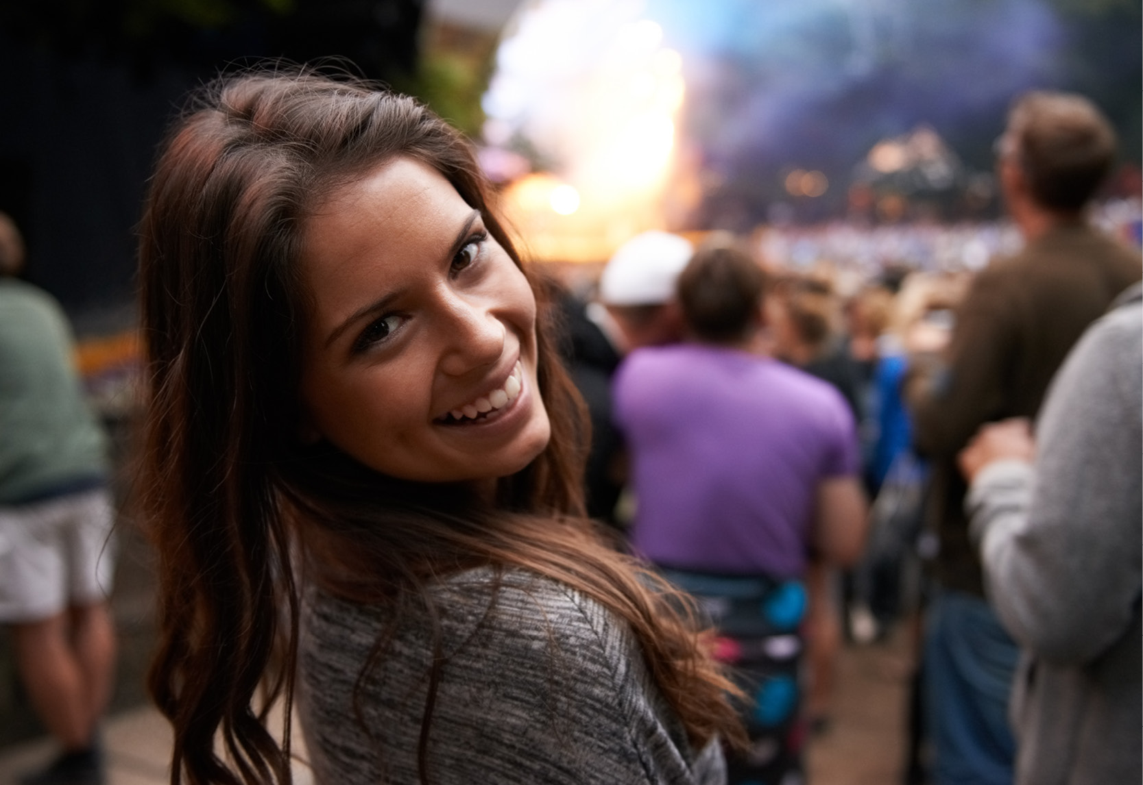 Une femme se tient dans un groupe de personnes lors d'un concert en plein air et regarde derrière elle par-dessus son épaule.