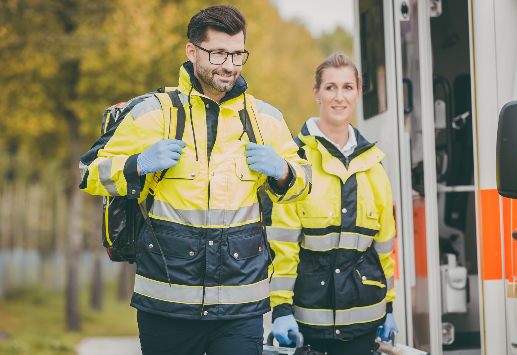 Un ambulancier avec un sac à dos et une ambulancière avec une trousse de premiers secours se tiennent devant leur véhicule d'intervention.