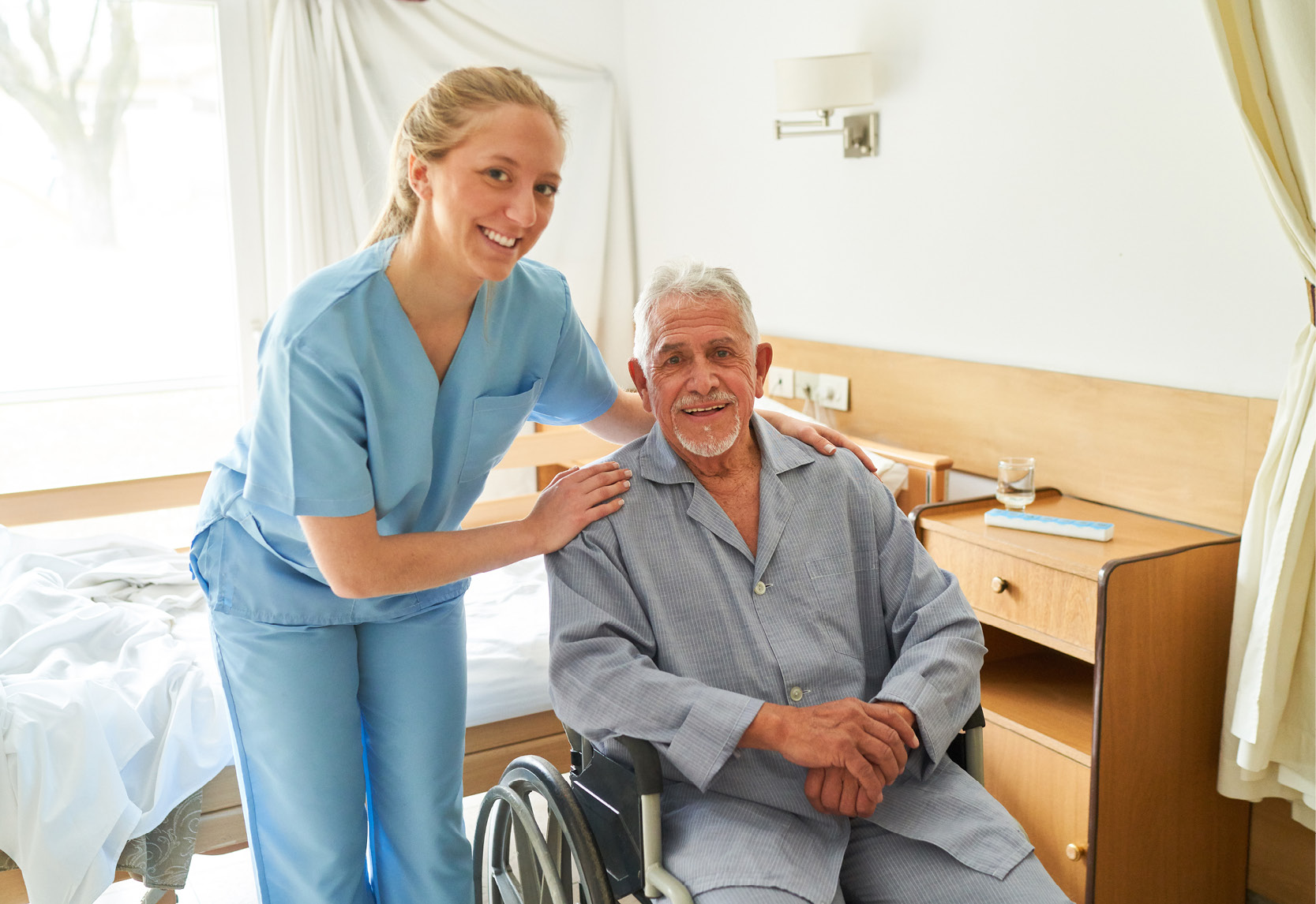 Une infirmière en tenue de travail bleu clair se tient à côté d'un patient en fauteuil roulant dans une chambre d'hôpital.